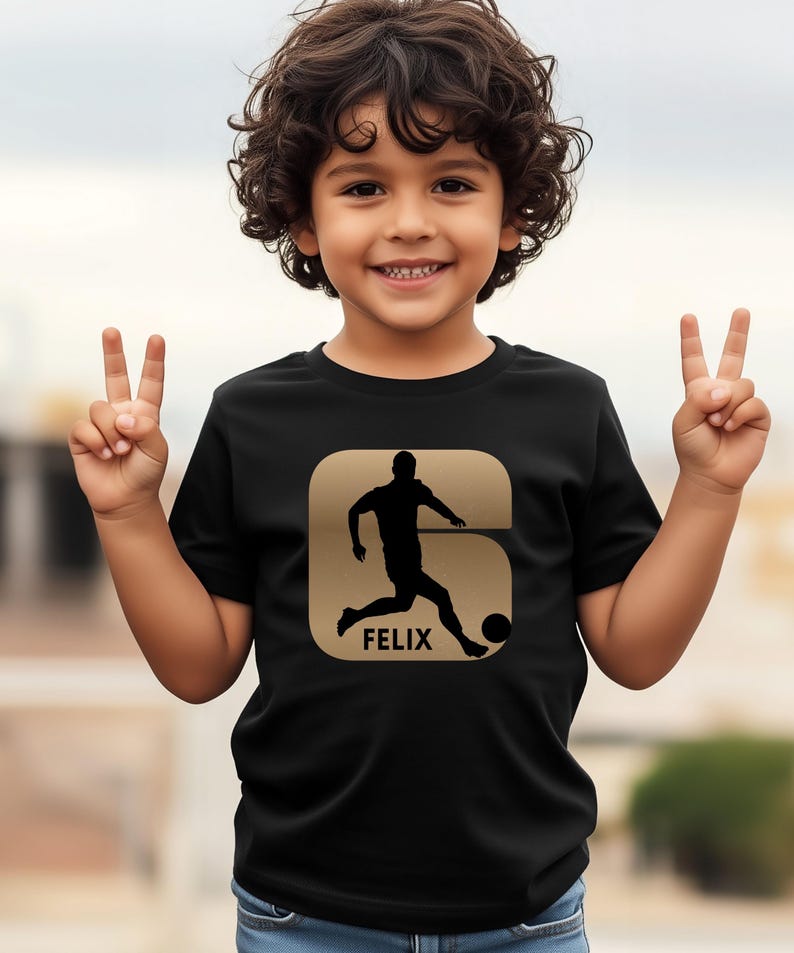 Pode incluir: Camiseta preta com um desenho dourado de um jogador de futebol chutando uma bola, com o nome "FELIX" abaixo. O desenho est&aacute; dentro de uma forma dourada. A camiseta &eacute; usada por uma crian&ccedil;a.