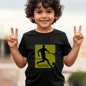 Pode incluir: Camiseta preta com a silhueta de um jogador de futebol dentro de um n&uacute;mero 5 verde. O nome "MATTEO" est&aacute; impresso abaixo. Uma crian&ccedil;a sorri e levanta dois dedos de cada m&atilde;o.