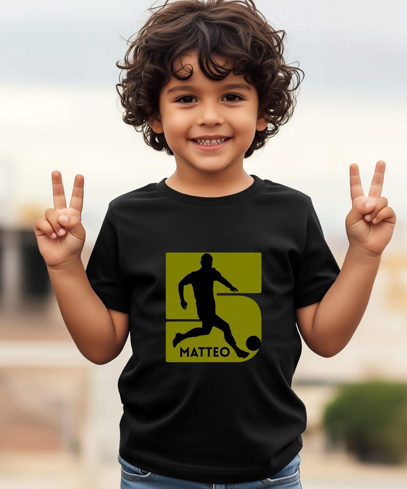 Pode incluir: Camiseta preta com a silhueta de um jogador de futebol dentro de um n&uacute;mero 5 verde. O nome "MATTEO" est&aacute; impresso abaixo. Uma crian&ccedil;a sorri e levanta dois dedos de cada m&atilde;o.