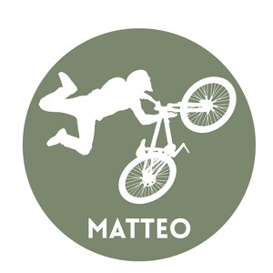May include: A circular olive green emblem featuring a white silhouette of a person performing a stunt on a mountain bike with one wheel elevated. The name MATTEO is printed in white capital letters at the bottom of the circular graphic design.