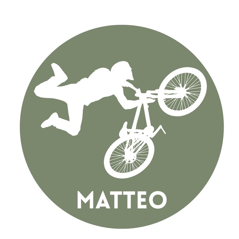 May include: A circular olive green emblem featuring a white silhouette of a person performing a stunt on a mountain bike with one wheel elevated. The name MATTEO is printed in white capital letters at the bottom of the circular graphic design.
