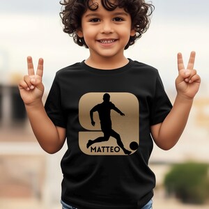 Pode incluir: Camiseta preta com um desenho dourado de um jogador de futebol chutando uma bola, com o nome "MATTEO" abaixo. A camiseta &eacute; usada por uma crian&ccedil;a que sorri e faz o sinal de vit&oacute;ria com as duas m&atilde;os.