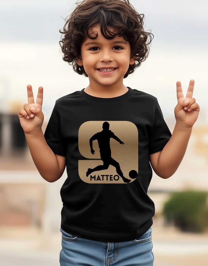 Pode incluir: Camiseta preta com um desenho dourado de um jogador de futebol chutando uma bola, com o nome "MATTEO" abaixo. A camiseta &eacute; usada por uma crian&ccedil;a que sorri e faz o sinal de vit&oacute;ria com as duas m&atilde;os.