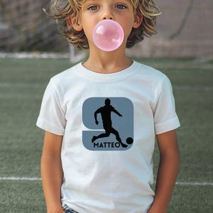 Puede incluir: Una camiseta blanca de manga corta con un gr&aacute;fico de la silueta negra de un jugador de f&uacute;tbol pateando un bal&oacute;n dentro de un cuadrado azul claro de esquinas redondeadas, con el nombre MATTEO impreso en letras may&uacute;sculas negras debajo.