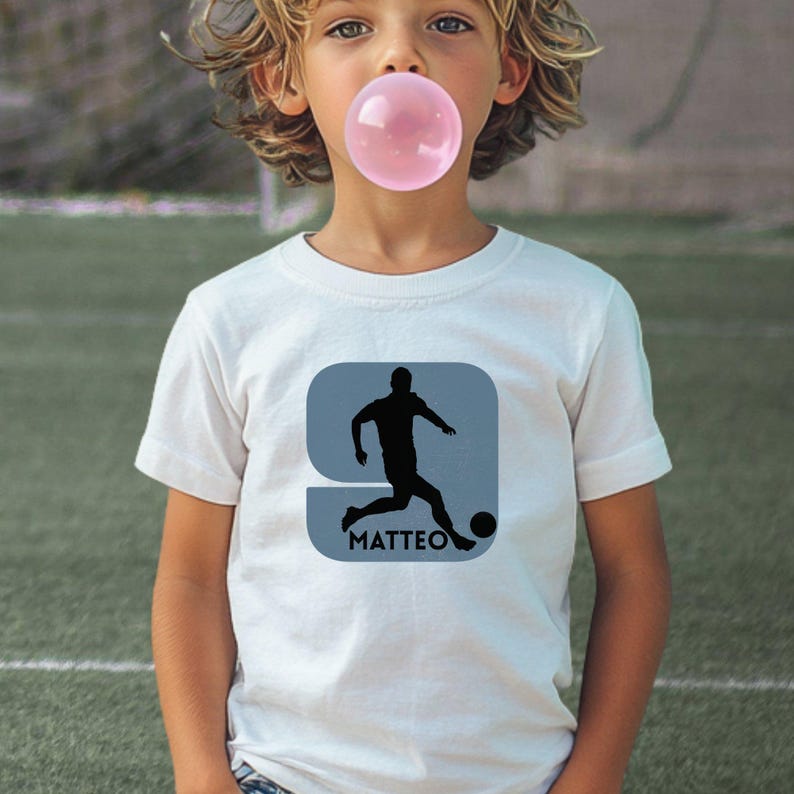Puede incluir: Una camiseta blanca de manga corta con un gr&aacute;fico de la silueta negra de un jugador de f&uacute;tbol pateando un bal&oacute;n dentro de un cuadrado azul claro de esquinas redondeadas, con el nombre MATTEO impreso en letras may&uacute;sculas negras debajo.