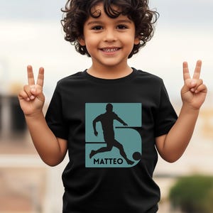 Pode incluir: Camiseta preta com um gr&aacute;fico azul-petr&oacute;leo de um jogador de futebol chutando uma bola, o n&uacute;mero 5 e o nome "MATTEO". A camiseta &eacute; usada por uma crian&ccedil;a pequena.