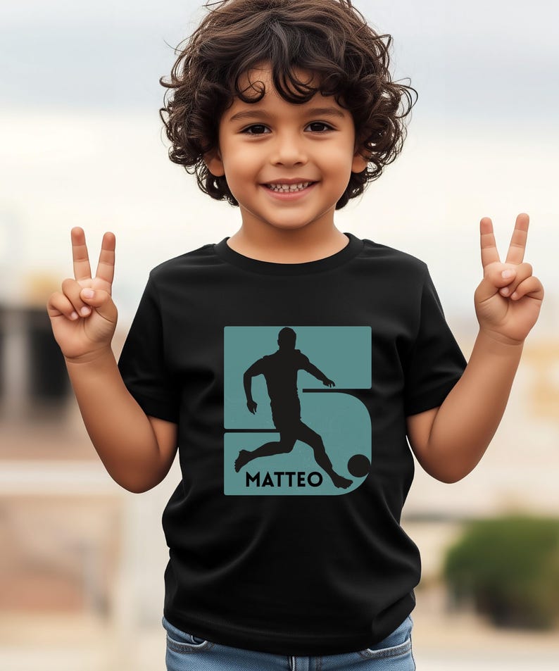 Pode incluir: Camiseta preta com um gr&aacute;fico azul-petr&oacute;leo de um jogador de futebol chutando uma bola, o n&uacute;mero 5 e o nome "MATTEO". A camiseta &eacute; usada por uma crian&ccedil;a pequena.