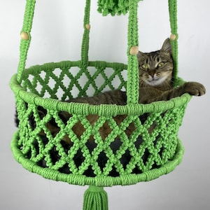 May include: A bright green macrame hanging cat bed featuring a circular base with a diamond-patterned woven side, suspended by four braided cords with small wooden beads, and finished with a decorative tassel hanging from the centre bottom.