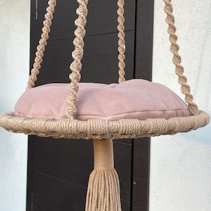 Puede incluir: Una cama colgante para gatos con un cojín redondo color rubor. La cama está suspendida por una gruesa cuerda beige trenzada. Una borla de cuerda a juego cuelga debajo del cojín. La cama está diseñada para uso en interiores.