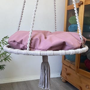 May include: A circular hanging swing chair featuring a dusty pink cushion set within a white macrame frame. The seat is suspended by four thick, twisted white rope cords and includes a decorative white tassel hanging from the center base.