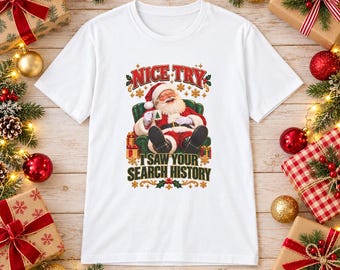 May include: A white short-sleeved t-shirt featuring a graphic of Santa Claus sitting in a green armchair holding a mug. The text above reads NICE TRY and the text below reads I SAW YOUR SEARCH HISTORY in red and dark green lettering.