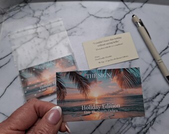 May include: A business card with a tropical sunset design, palm trees, and the text "THE SIGN Holiday Edition Etsy TheSignByDebra." A clear plastic bag contains a similar card. A pen and a small card with text are also present.