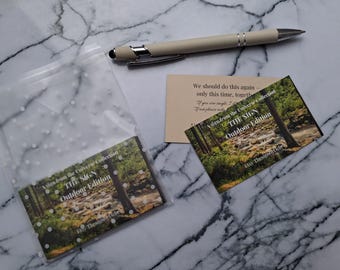 May include: An ivory pen rests near two cards and a clear plastic bag with a white dot pattern. The cards feature a forest scene and text that reads "THE SIGN Outdoor Edition" and "We should do this again..."