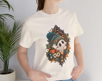 May include: A cream-colored short-sleeve t-shirt featuring a graphic of a white ghost wearing a teal knit hat, peeking through a zippered opening. The design includes autumn leaves, a small orange pumpkin, and the word Boo in teal.