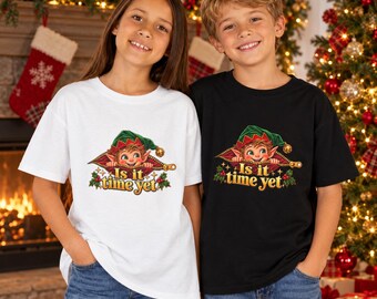 May include: A white t-shirt and a black t-shirt, each featuring a graphic of an elf peeking out from behind a zipper. Below the elf, the text reads "Is it time yet" in gold lettering, accented with small green holly leaves and red berries.