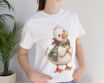 May include: A white short-sleeved t-shirt featuring a graphic illustration of a fluffy white duckling wearing a green and white striped scarf with holly berries and holding a gold star. The duckling has orange feet and an orange beak.