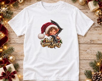 May include: A white t-shirt featuring a graphic of a cartoon elf wearing a red Santa hat and peeking out from a zipper opening. Below the elf, the text reads Is it time yet? in a decorative gold font with small holly berry accents.