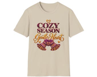 May include: A beige short-sleeved t-shirt featuring a graphic print with the text COZY SEASON and Gentle Heart in dark red and gold lettering. The design includes a purple and orange knit scarf illustration and small gold hearts.