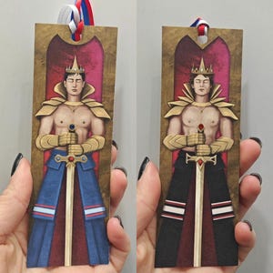 Könnte beinhalten: Zwei Lesezeichen mit Illustrationen einer gekrönten Figur, die ein Schwert hält. Eines hat ein blaues und rotes Design, das andere ein schwarzes und rotes Design. Beide haben goldene Akzente und ein Band zum Markieren von Seiten. Jedes Lesezeichen ist etwa 15 cm lang.