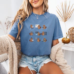 May include: A blue t-shirt with a repeating pattern of cartoon beavers engaged in tech activities. The beavers are shown with computers and coding symbols. The shirt is a solid blue colour and has a loose fit.
