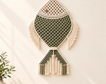 May include: A macrame wall hanging shaped like a fish, crafted from cream and olive green cord. The design features a knotted lattice pattern, fringed fins, and a tail with hanging tassels. A single dark bead serves as the fish eye.