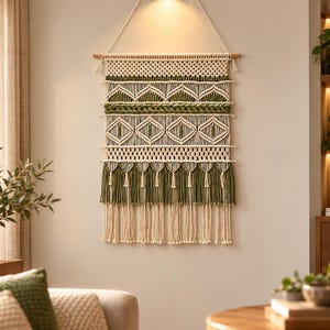 May include: A macrame wall hanging featuring off-white and olive green cotton cord. The design includes intricate knotted patterns, horizontal braided sections, diamond-shaped motifs, and a bottom fringe with alternating green and white tassels.