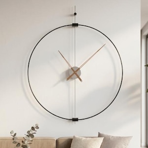 Peut inclure: Une grande horloge murale moderne au design minimaliste. L'horloge présente un cadre en métal noir formant un grand cercle, avec de fines aiguilles marron clair. Le cadran de l'horloge est de couleur gris clair. L'horloge est fixée sur un mur blanc.