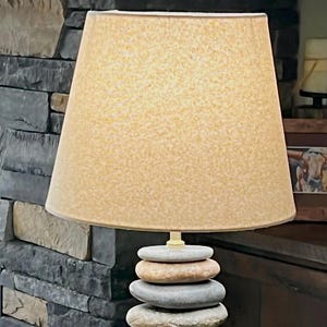 May include: A decorative lamp with a beige lampshade. The base is a stack of smooth, rounded stones in shades of gray and tan. The lamp is on a wooden surface, with a stone wall in the background.