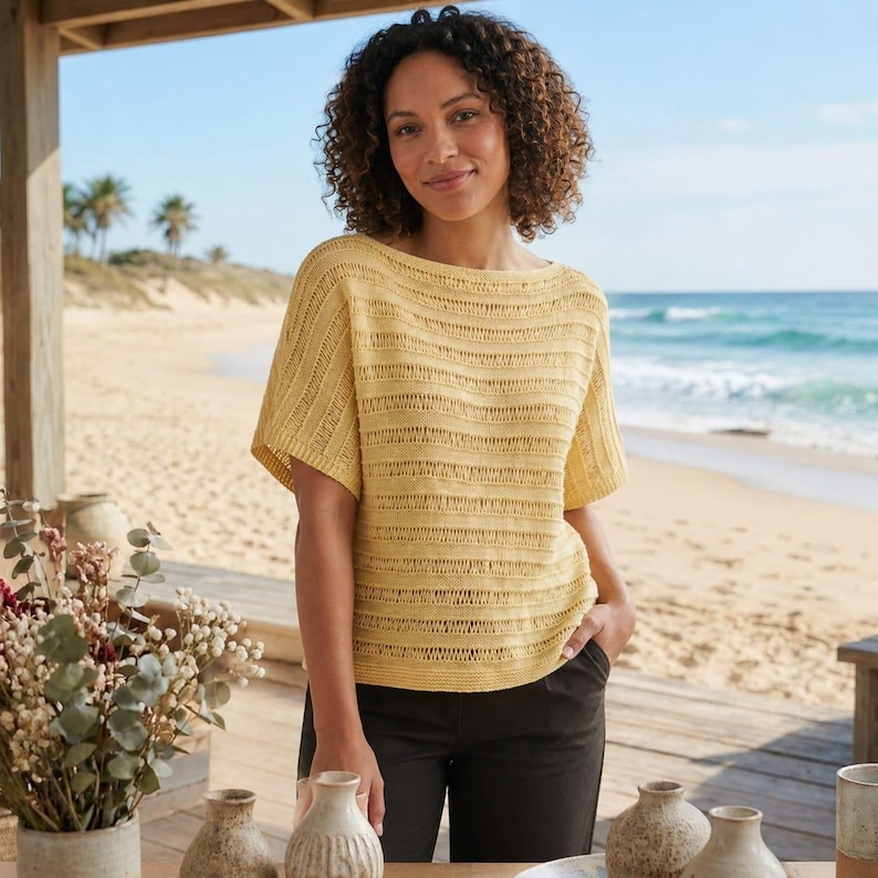 May include: A woman wearing a short-sleeved, yellow knit top with horizontal striped detailing. The top has a boat neck and is paired with dark pants. The background features a beach scene with the ocean and palm trees.