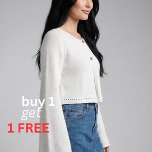 May include: A white, cropped cardigan with bell sleeves and button closures. The model is wearing a denim skirt. The text "buy 1 get 1 FREE" is displayed in white and red.