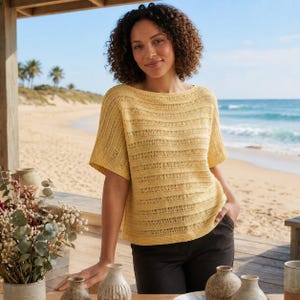 May include: A yellow short-sleeved top with a boat neck. The top has a horizontal striped pattern. The person is wearing black pants. The background shows a beach with palm trees and the ocean.