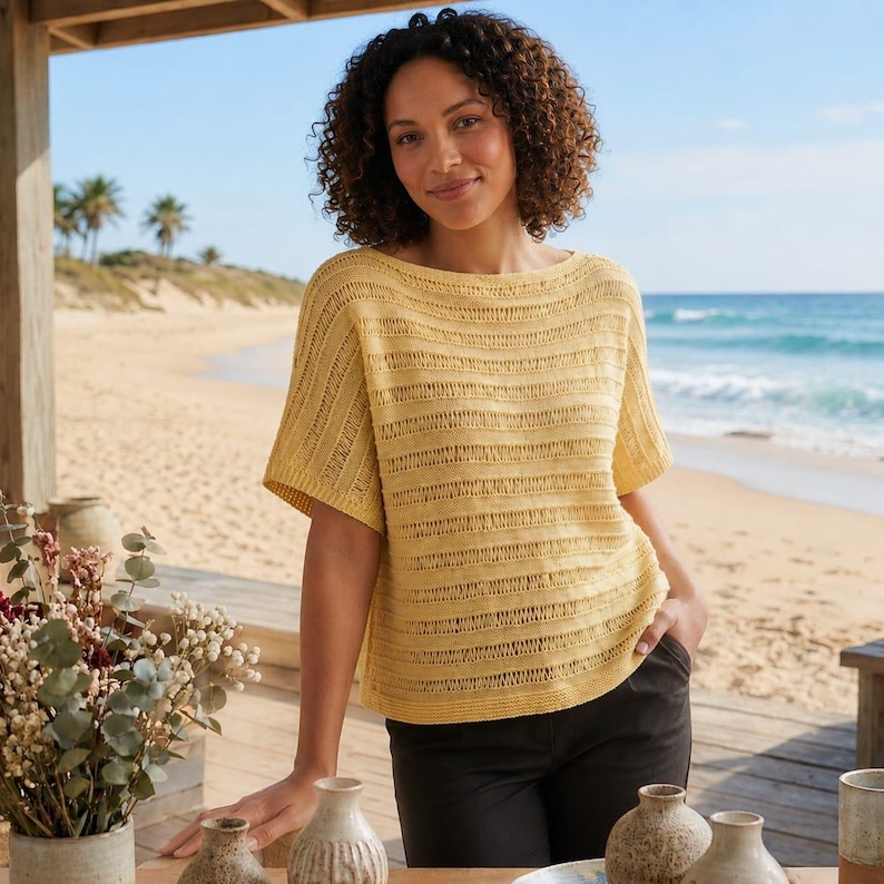 May include: A yellow short-sleeved top with a boat neck. The top has a horizontal striped pattern. The person is wearing black pants. The background shows a beach with palm trees and the ocean.