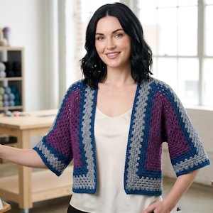 May include: A handmade, open-front crochet cardigan in a granny square pattern. The short-sleeved garment features a color block design with shades of purple, gray, and blue. The cardigan is worn over a white top.