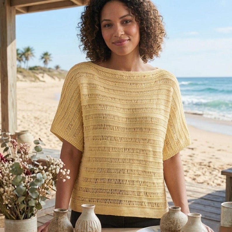 May include: A woman wearing a short-sleeved, yellow knit top with a boat neck. The top has a horizontal striped pattern. The background shows a beach with the ocean and palm trees.
