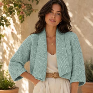 May include: A light mint green open-front cardigan featuring a textured knit pattern, three-quarter length sleeves, and a wide shawl collar. The garment is worn over a cream-coloured top with a tan belt, and a simple gold necklace is visible.