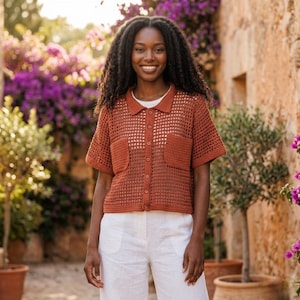 Puede incluir: Una camisa de crochet de manga corta, color óxido, con dos bolsillos delanteros y cuello. La camisa está abotonada y se lleva sobre una camiseta interior blanca. El modelo lleva pantalones blancos.