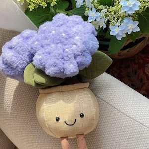 May include: A soft toy of a potted hydrangea. The toy has a light purple flower with white spots, green leaves, and a beige pot with a smiling face and small legs. Real flowers are in the background.