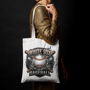 Op de afbeelding: Witte draagtas met een honkbalontwerp. Het ontwerp bevat een honkbal met rode stiksels, gekruiste knuppels en de tekst "WHITE SOX BASEBALL". De tas wordt vastgehouden door een persoon die een goudkleurige top draagt.