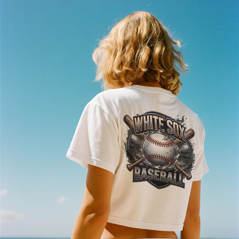 Op de afbeelding: Witte crop top met een honkbalafbeelding op de rug. De afbeelding toont een honkbal, knuppels en de woorden "WHITE SOX BASEBALL" in een schildontwerp. Het shirt wordt gedragen tegen een blauwe lucht.