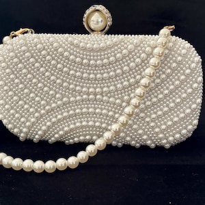 Pode incluir: Uma clutch retangular branca coberta de pérolas. Possui uma alça de pérolas e um fecho decorativo com uma pérola grande. Um design elegante, ideal para eventos formais e casamentos.