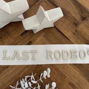 Könnte beinhalten: Eine weiße Satinschärpe mit dem Schriftzug LAST RODEO in perlenbesetzten Buchstaben, flankiert von zwei mit Perlen verzierten Herzformen. Die Schärpe liegt neben zwei weißen sternförmigen Ornamenten und einem kleinen weißen Kunstzweig.