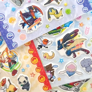 Puede incluir: Hojas de pegatinas Pokémon coloridas con varios personajes. Las pegatinas tienen varias formas y tamaños, algunas con la etiqueta "MEGA" y nombres de personajes. Las pegatinas están en hojas de plástico transparente con bordes decorativos y estrellas.