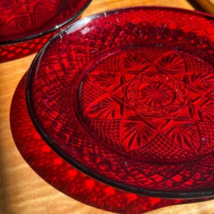 May include: Two ruby red glass plates with intricate starburst and geometric patterns. The plates are on a wooden surface, with sunlight casting shadows. The plates have a dark rim and a detailed, textured design.