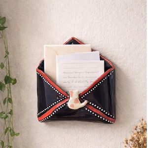 Könnte beinhalten: Schwarzer, wandmontierter Briefhalter aus Keramik in Umschlagform mit rotem Rand und weißen Punkten. Eine kleine, weiße Katze mit rosa Flecken verziert die Vorderseite. Der Halter enthält Briefe.