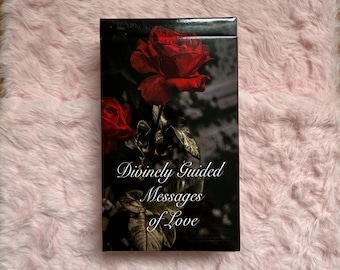 Puede incluir: Una caja rectangular con el texto "Divinely Guided Messages of Love" presenta un fondo oscuro con una gran rosa roja y rosas más pequeñas. La caja está sobre una superficie rosa y esponjosa.