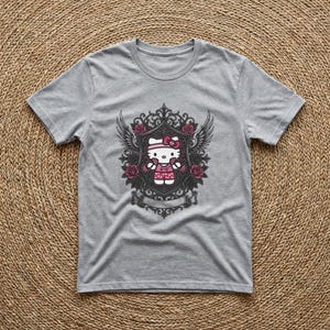 Può includere: T-shirt grigio con grafica di Hello Kitty. Il design presenta Hello Kitty con un vestito e fiocco rosa, circondata da rose, ali e una cornice decorativa. La grafica è centrata.