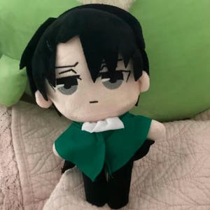 Peut inclure: Poupée en peluche d'un personnage aux cheveux noirs et aux yeux gris, portant une cape verte et un pantalon noir. La poupée a une expression neutre et est posée sur une surface à motifs. L'objet est un objet de collection.