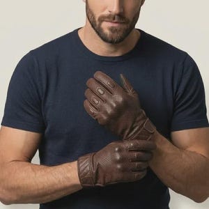 Könnte beinhalten: Ein Paar dunkelbraune Lederhandschuhe mit perforierter Textur an den Fingern und dem Handrücken. Die Handschuhe verfügen über eine verstärkte Knöchelpolsterung und einen sicheren Handgelenkverschluss mit einem gesteppten Muster am Bündchen.
