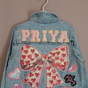Könnte beinhalten: Eine hellblaue Jeansjacke mit dem Namen PRIYA in rosa Buchstaben mit Goldrand. Die Rückseite ist mit einer großen Pailletten-Schleife mit roten Herzen, einem Herz-Aufnäher, einer Kamera, einer Wassermelone, dem Text girl gang und einer Sonnenbrille verziert.