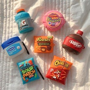 Op de afbeelding: Een verzameling van zeven siliconen beschermhoesjes voor draadloze oordopjes, met ontwerpen geïnspireerd op Gatorade, Hubba Bubba, Vaseline, Reese's, Dr. Pepper, Sour Patch Kids en Cheetos. Elk hoesje toont het bijbehorende merklogo en kleuren.